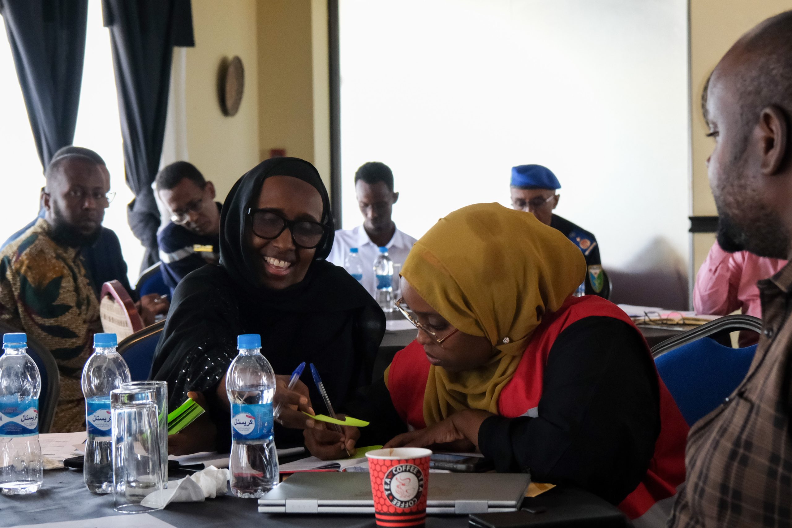 Several people sit around a table with water bottles, two women in the foreground discussing documents, one smiling and
