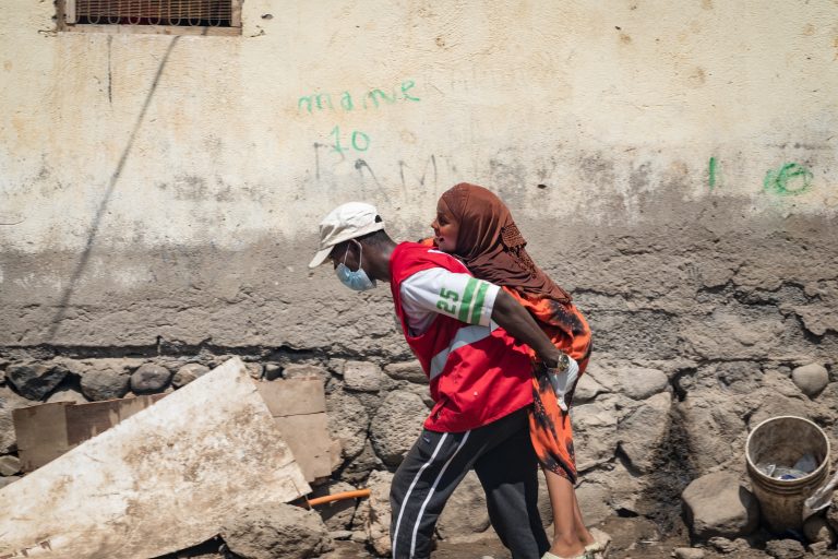 A person in a red vest and face mask carries another person on their back along a rough, debris-strewn street next to a