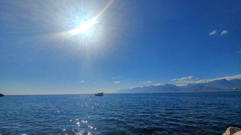 The sun shines brightly over a calm blue sea with a boat in the distance and mountains visible on the horizon under a cl