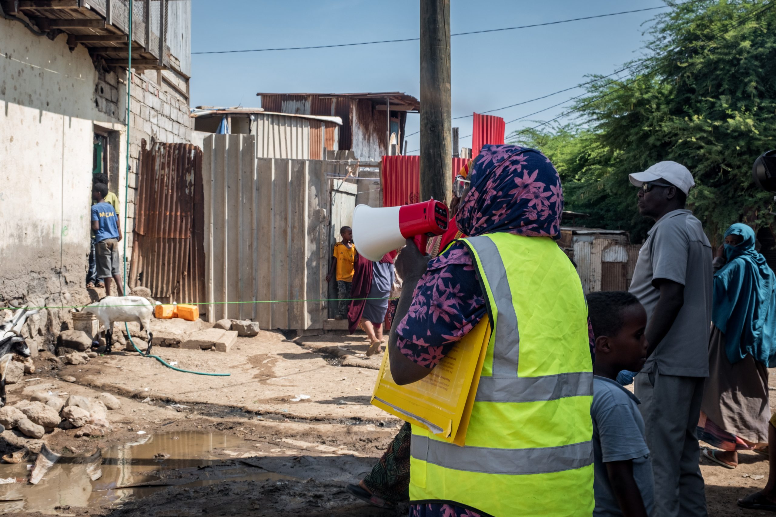 A person in a yellow vest holds a megaphone and documents while addressing people in an informal settlement area with sc