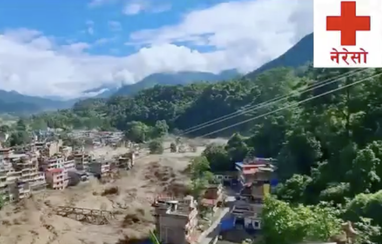 A flooded river runs through a town in a mountainous area; damaged buildings and debris are visible. A red cross with Ne