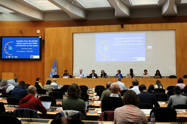 Panelists sit at a long table addressing an audience in a conference room, with a blue presentation slide displayed on l