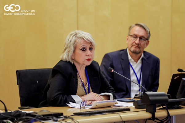 Two people sit at a conference table with microphones and laptops; one woman is speaking while the man listens. A “GEO G