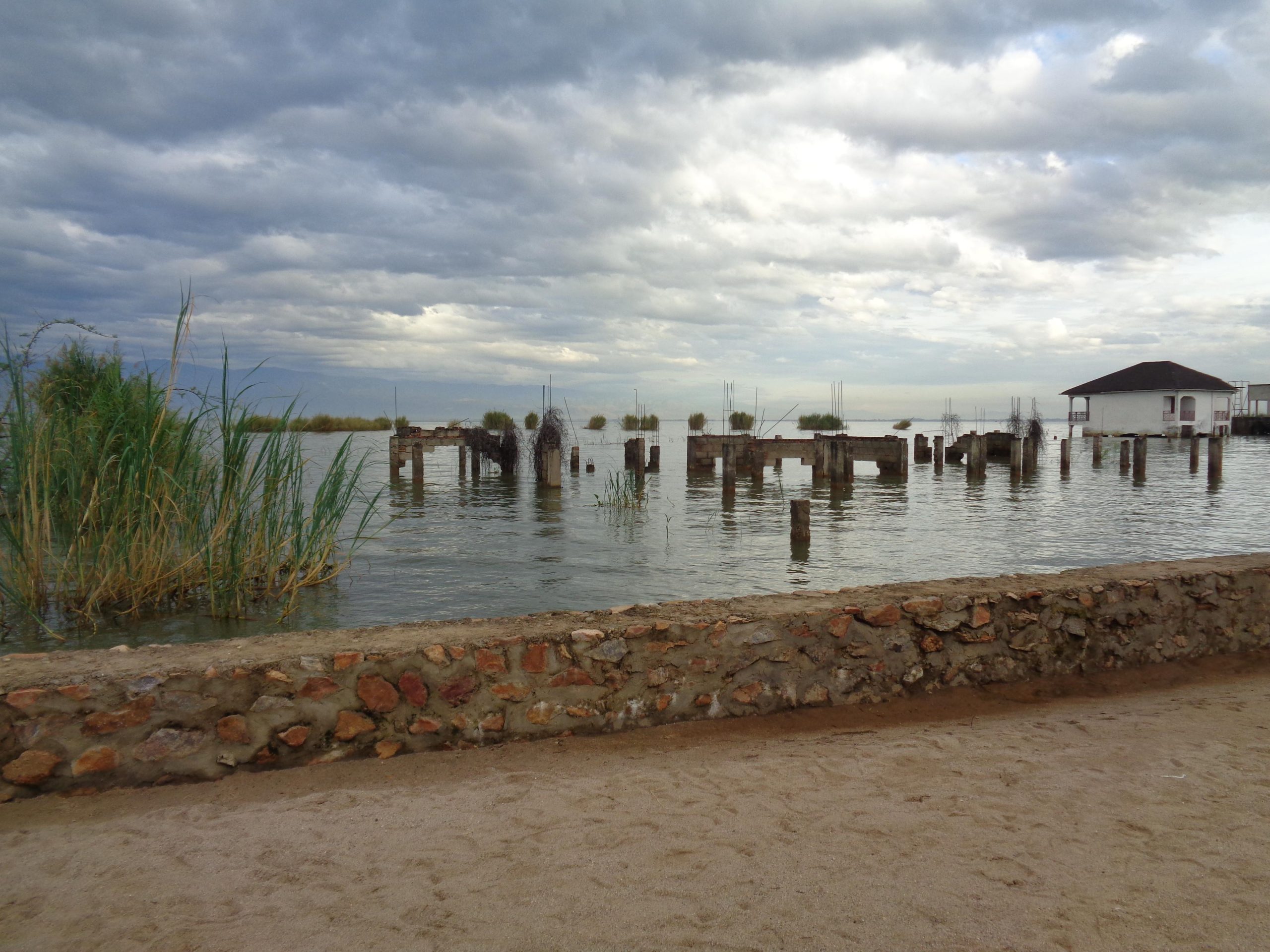 Sandy path with a stone wall, water with old wooden pilings, reeds on the left, and a small white building on the right