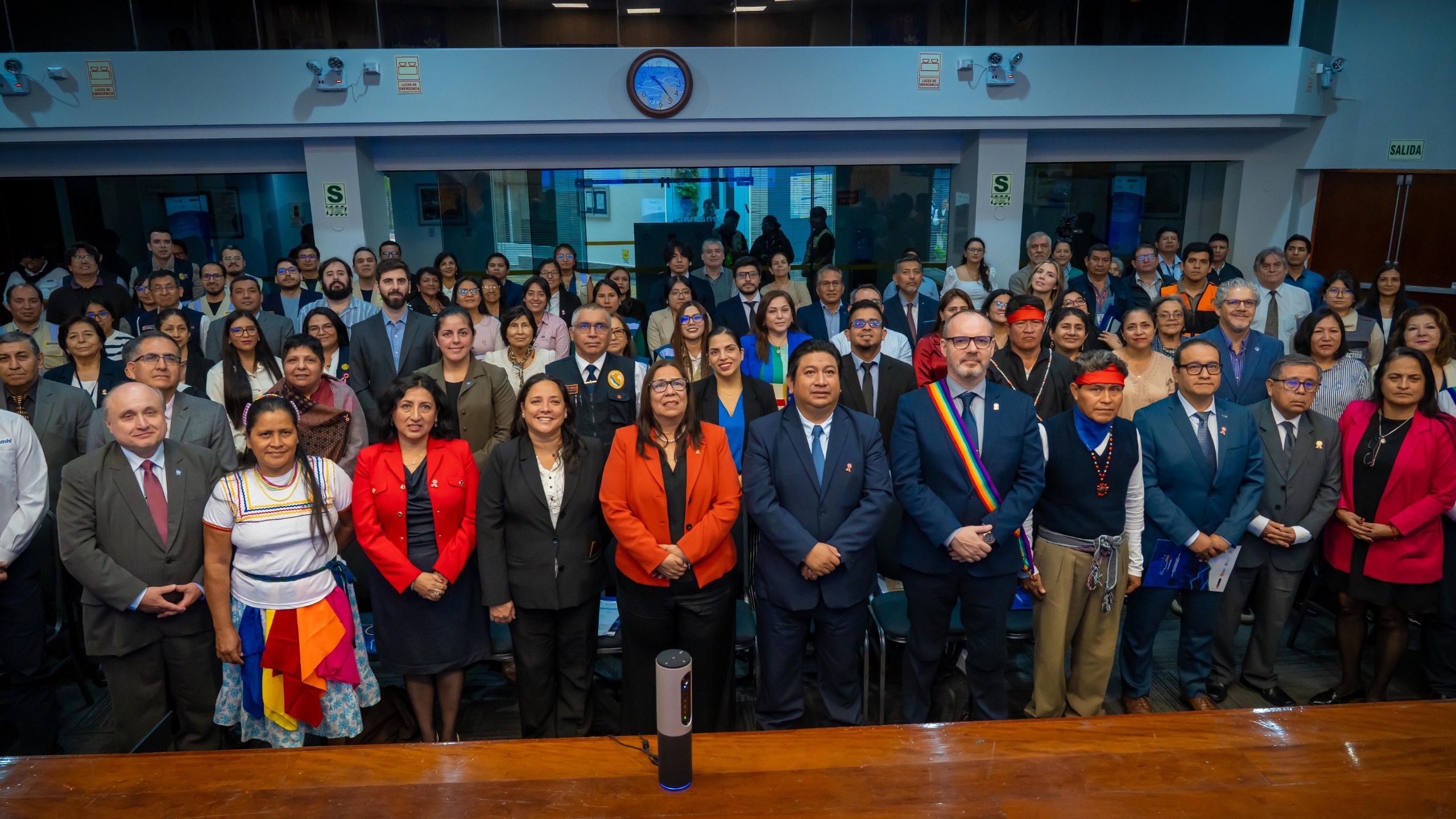 A large group of people, including men and women in formal and traditional attire, stand together in a conference room,