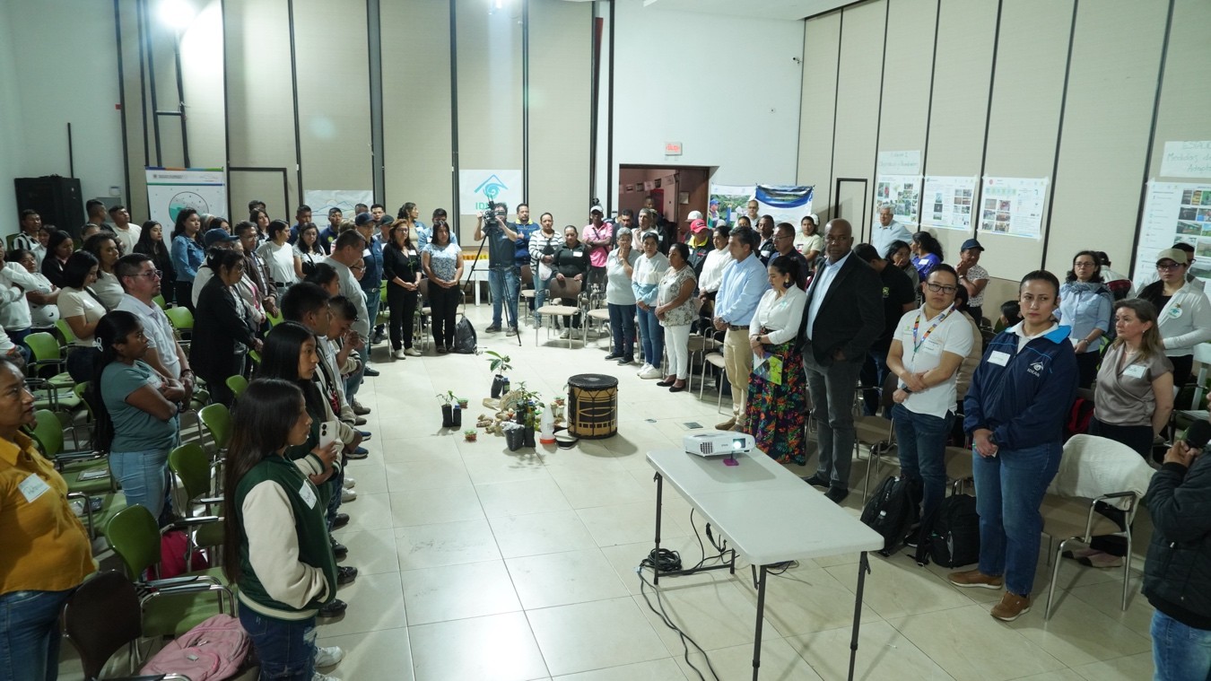 A large group of people stands in a conference room, facing each other in a circle around a display of plants and object