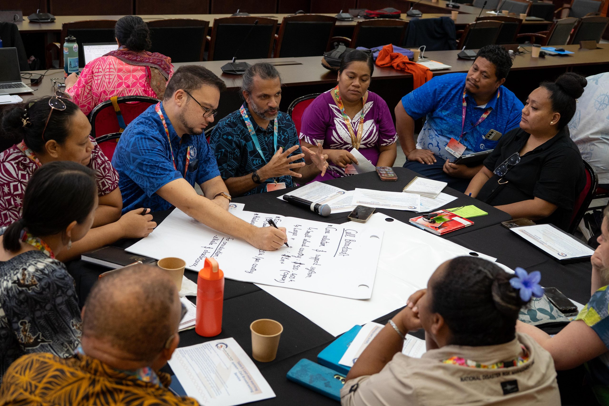 A group of people sit around a table in discussion, with one person writing on a large sheet of paper and documents, cup