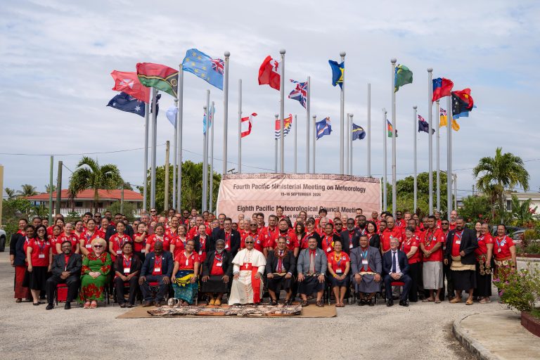 A large group of people pose for a photo in front of international flags and a banner for the Pacific Meteorological mee