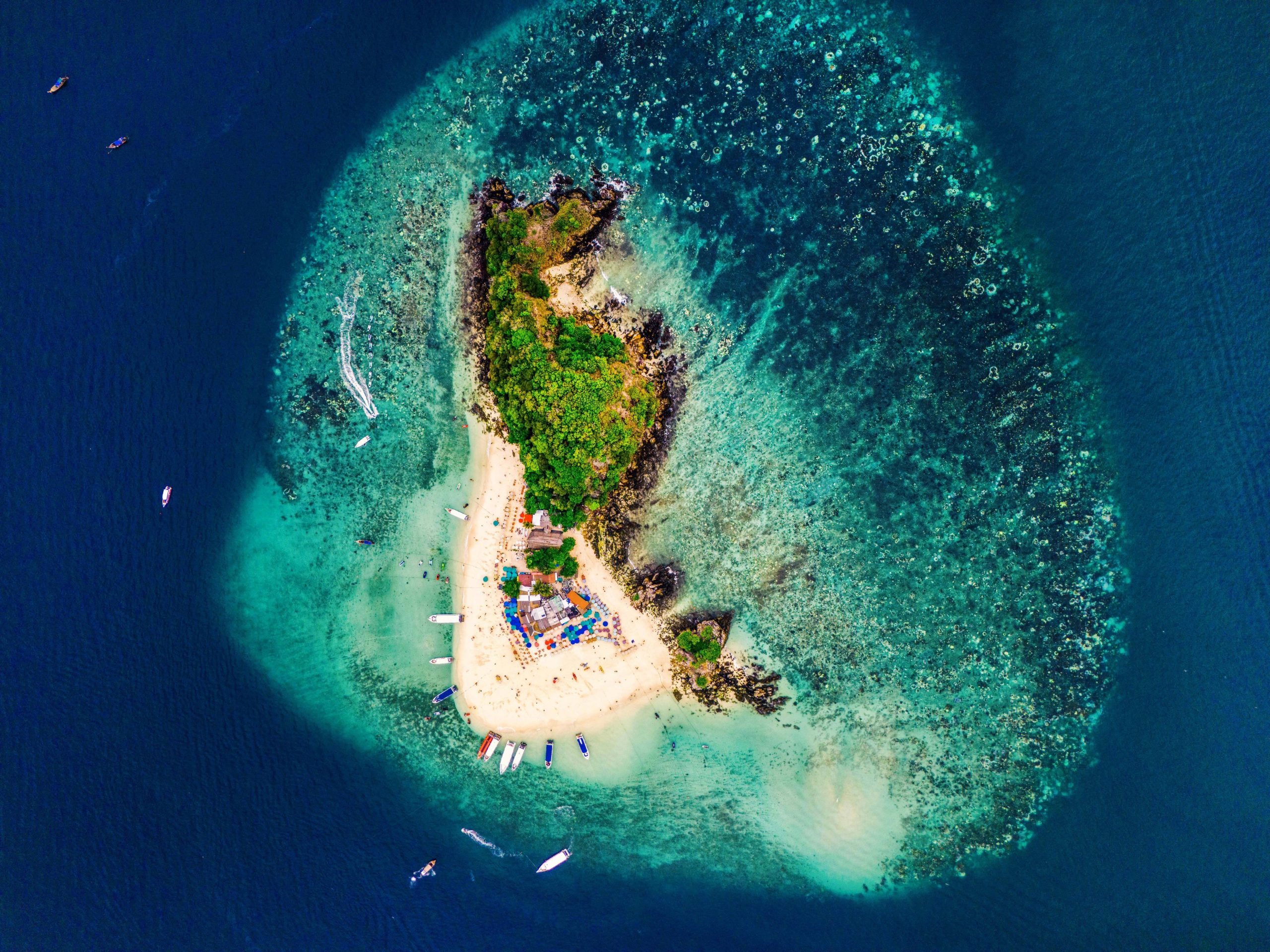 Aerial view of a small, lush island surrounded by a shallow coral reef and anchored boats.
