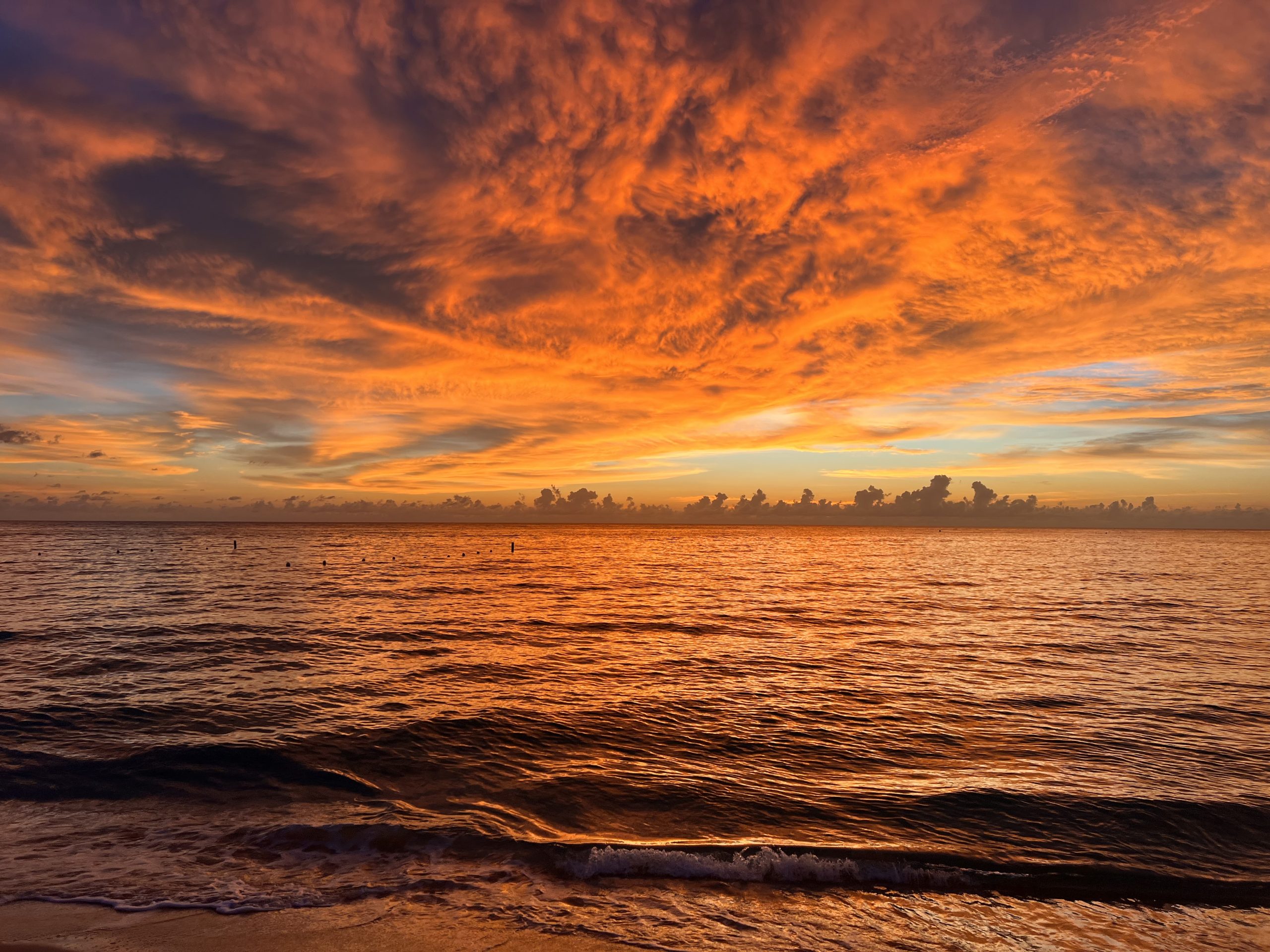 Orange and gold clouds fill the sky over a calm ocean at sunset, reflecting warm colors on the water and gentle waves ne