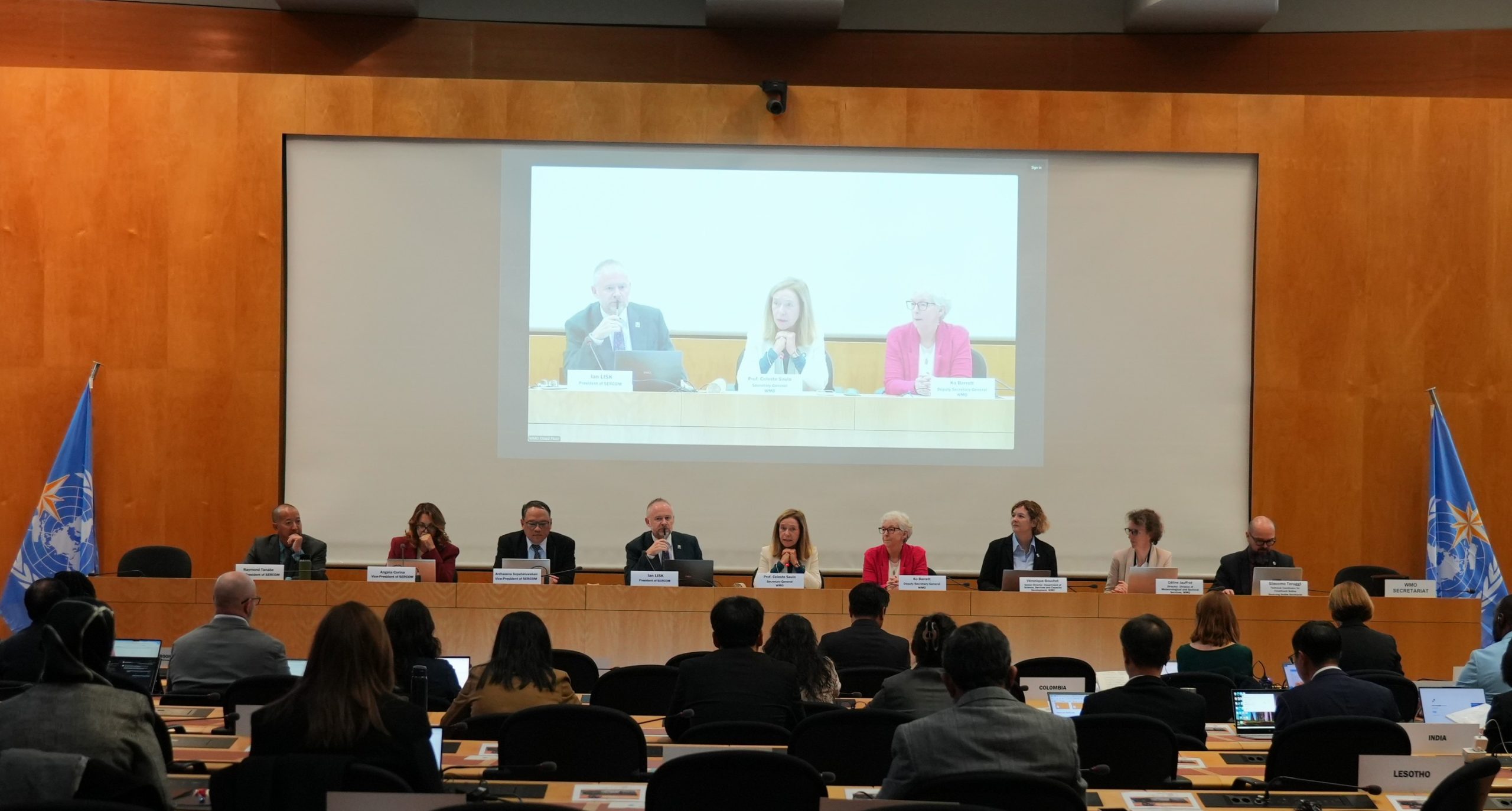 A large conference room with delegates seated at tables facing a projection screen displaying three people participating
