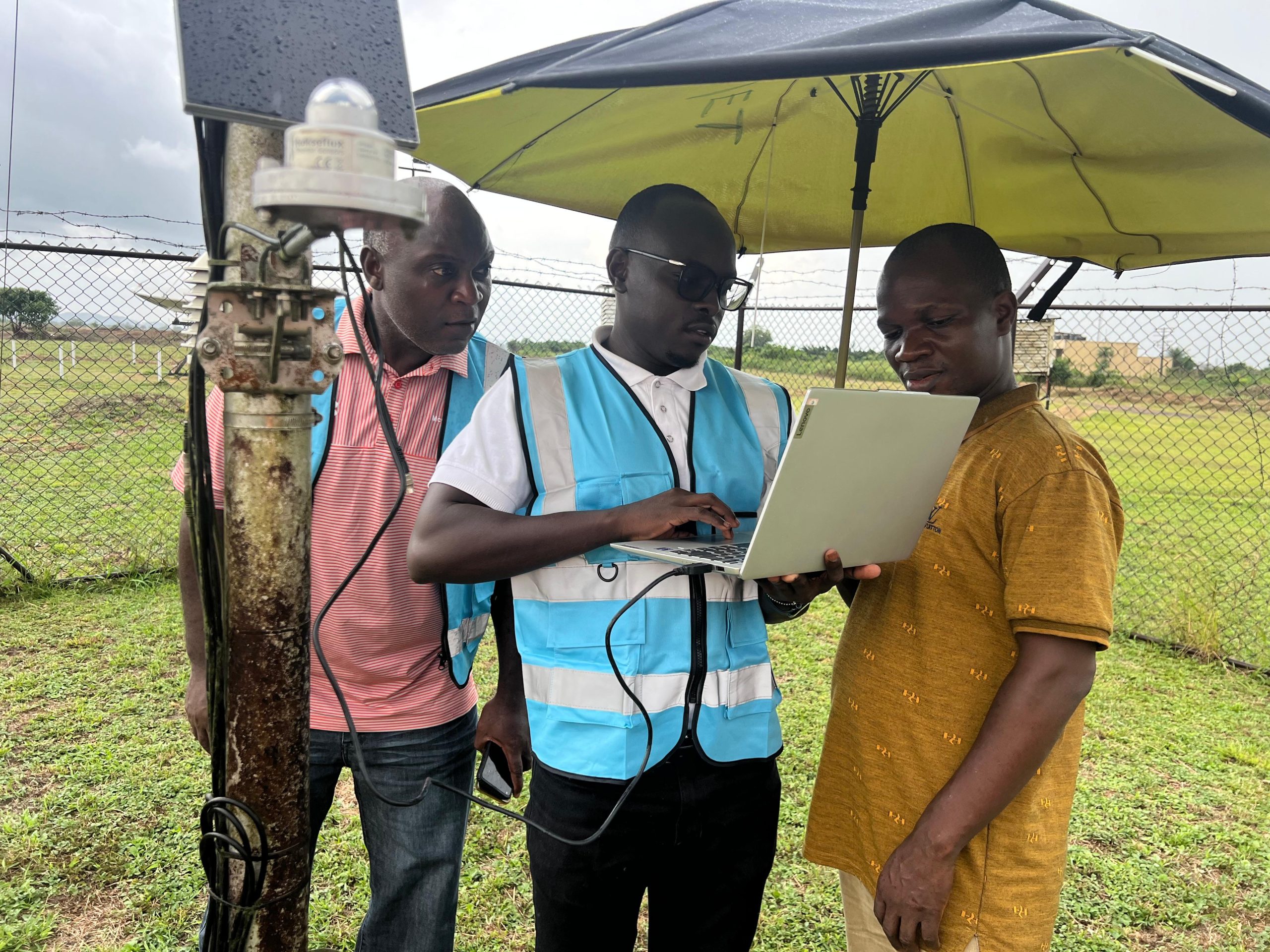 Three men stand outdoors beside meteorological equipment; one in a blue vest uses a laptop under a large umbrella, while